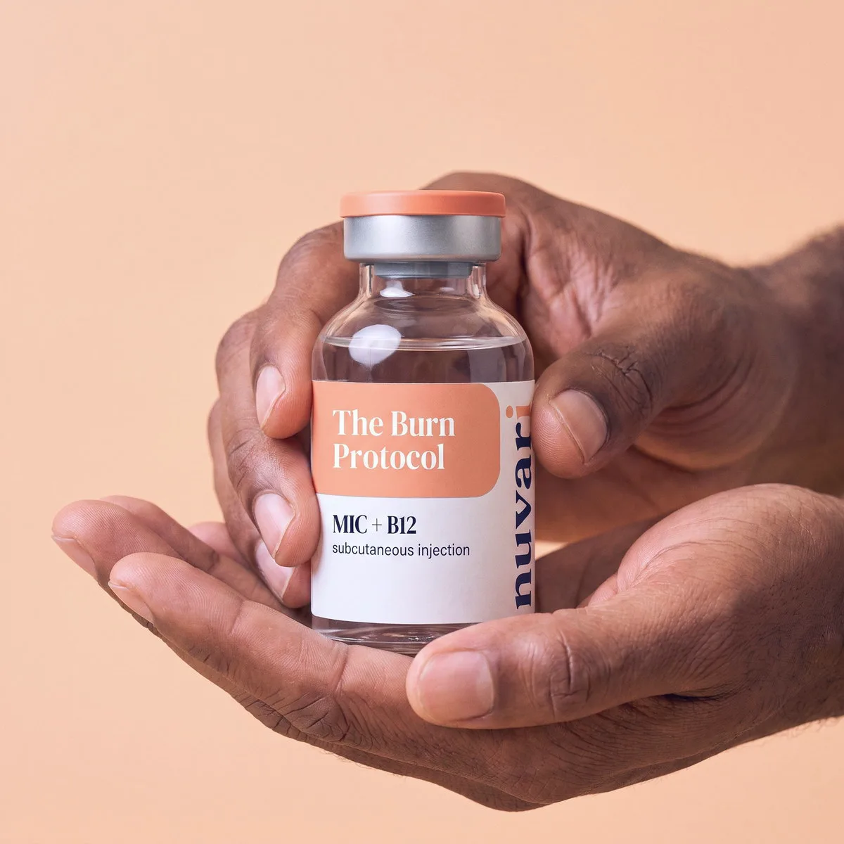 Two hands holding The Burn Protocol MIC + B12 injection vial, fingers curled around the glass, on a peach studio backdrop