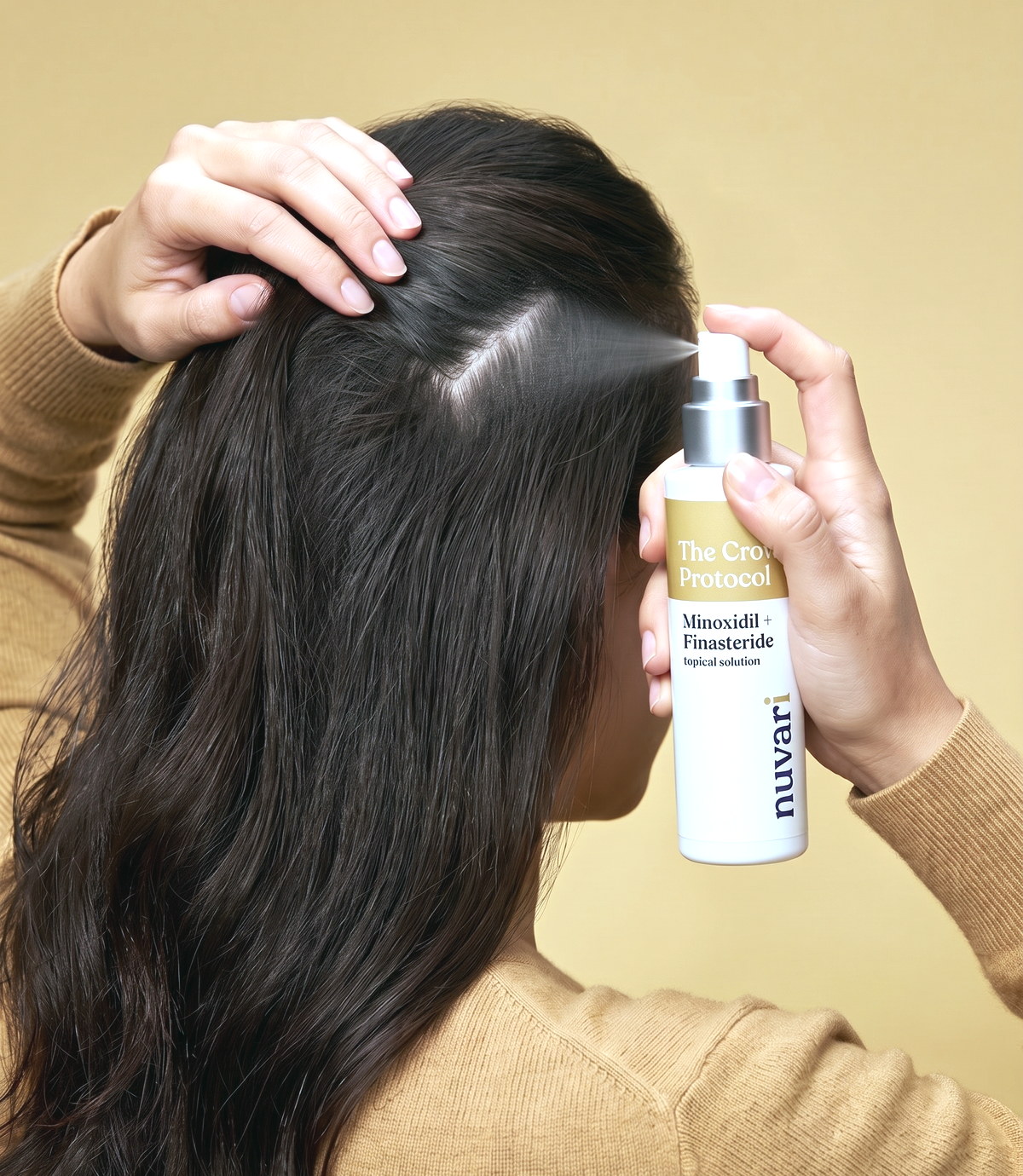 A woman seen from behind in a camel knit, holding her hair open at the crown parting and spraying the CROWN Protocol topical solution onto her scalp, on a warm gold backdrop