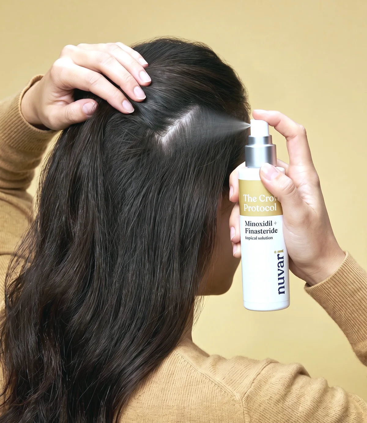 A woman seen from behind in a camel knit, holding her hair open at the crown parting and spraying the CROWN Protocol topical solution onto her scalp, on a warm gold backdrop