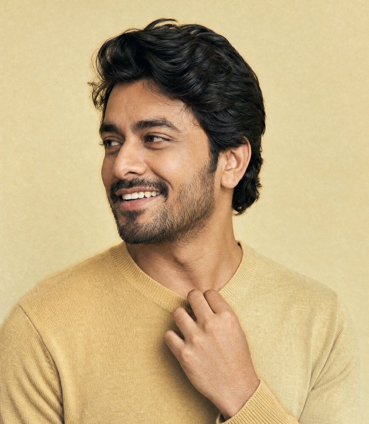 A smiling man in a camel knit with thick dark hair, glancing off to one side, one hand resting at his collar, on a warm gold backdrop