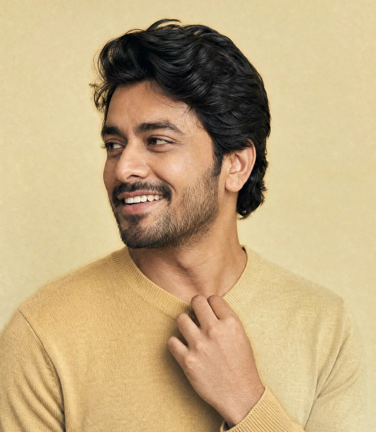 A smiling man in a camel knit with thick dark hair, glancing off to one side, one hand resting at his collar, on a warm gold backdrop