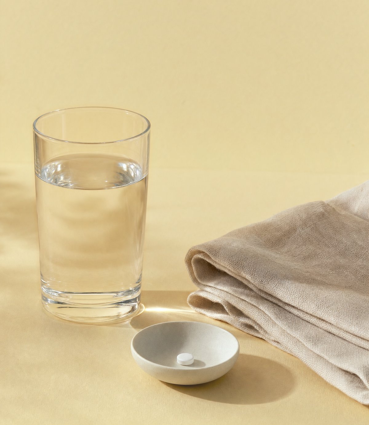 Morning still life: a glass of water, a folded linen cloth and a small ceramic dish holding one tablet on a warm gold surface