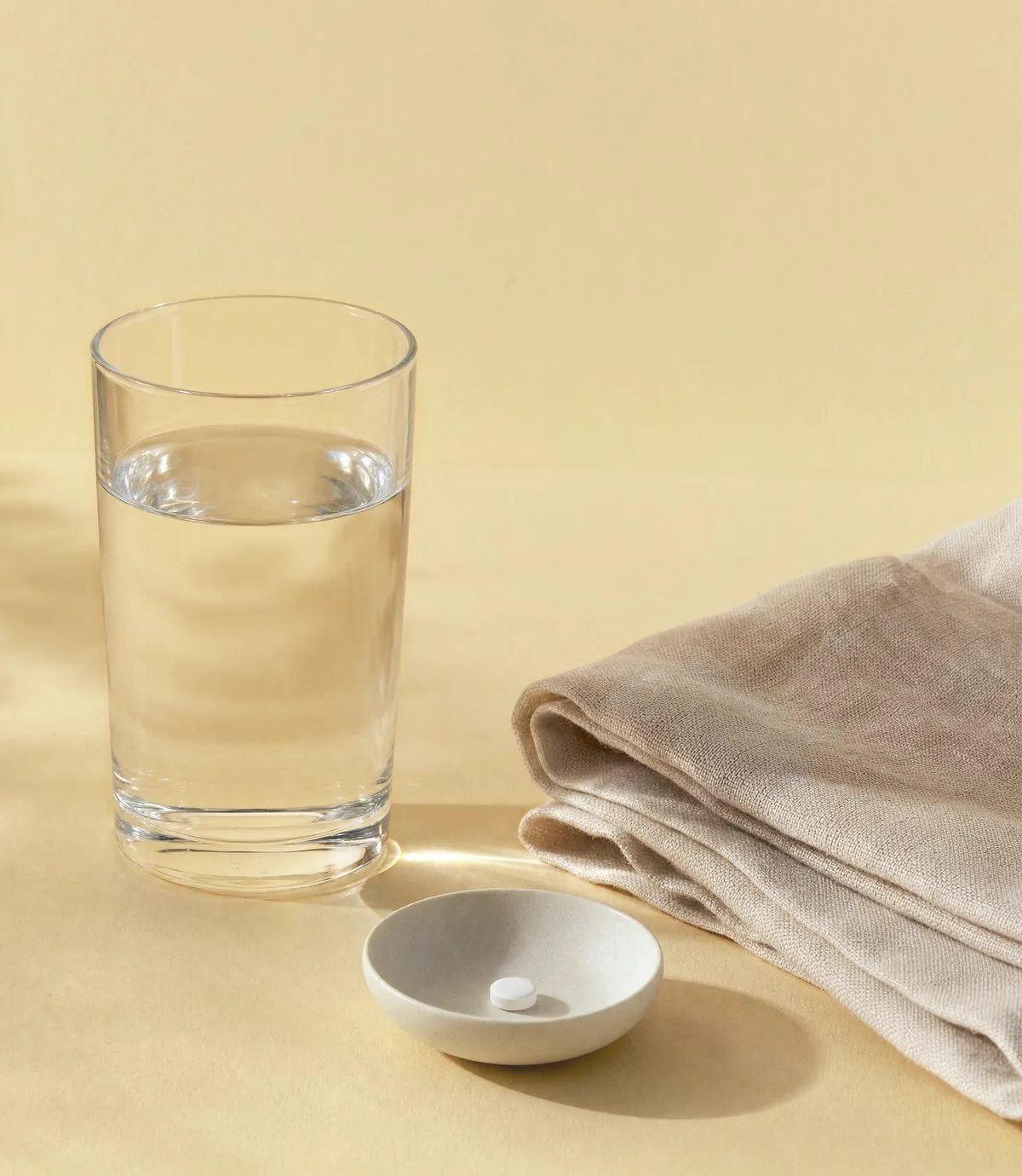 Morning still life: a glass of water, a folded linen cloth and a small ceramic dish holding one tablet on a warm gold surface