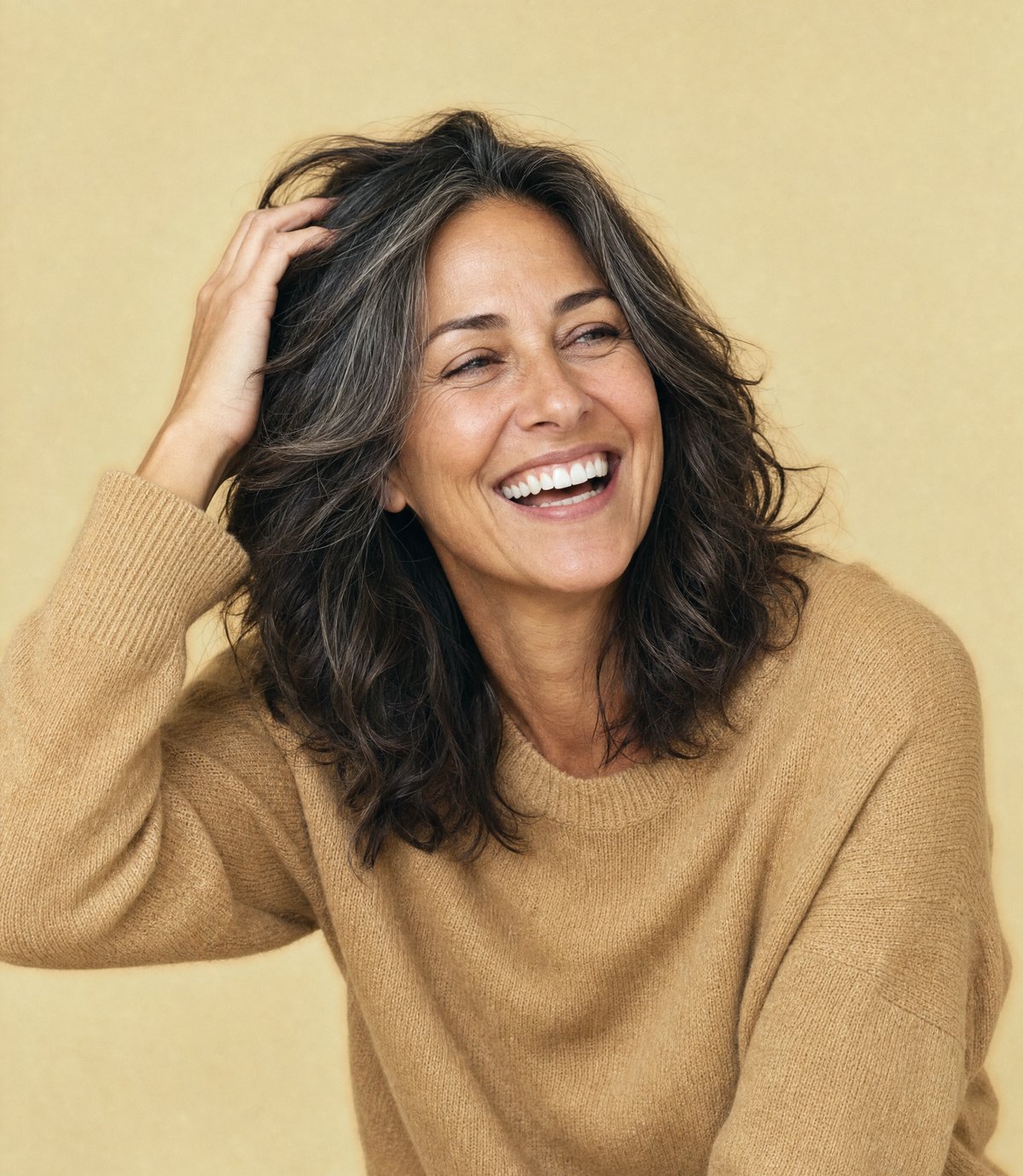 A woman in her mid-forties laughing as she runs a hand through thick shoulder-length hair on a warm gold backdrop