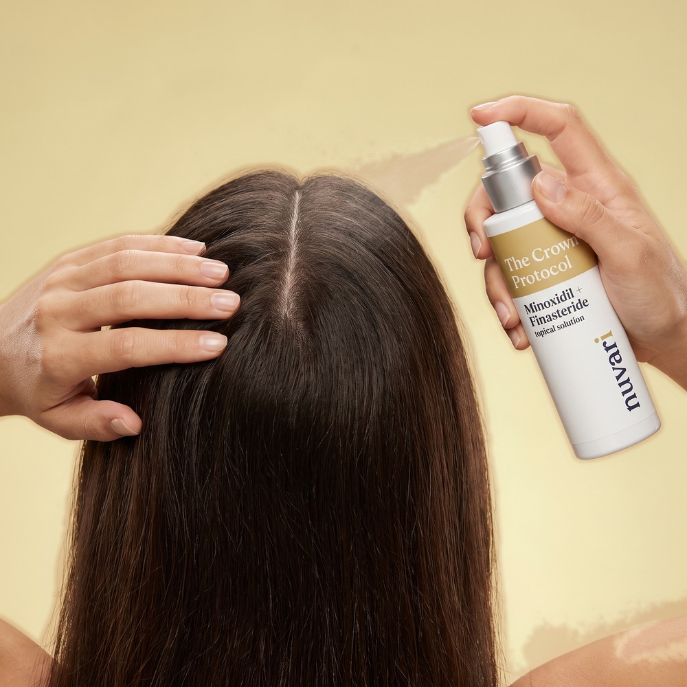 A woman holding the CROWN Protocol topical solution bottle upright and spraying it along the part in her dark hair, on a warm gold backdrop