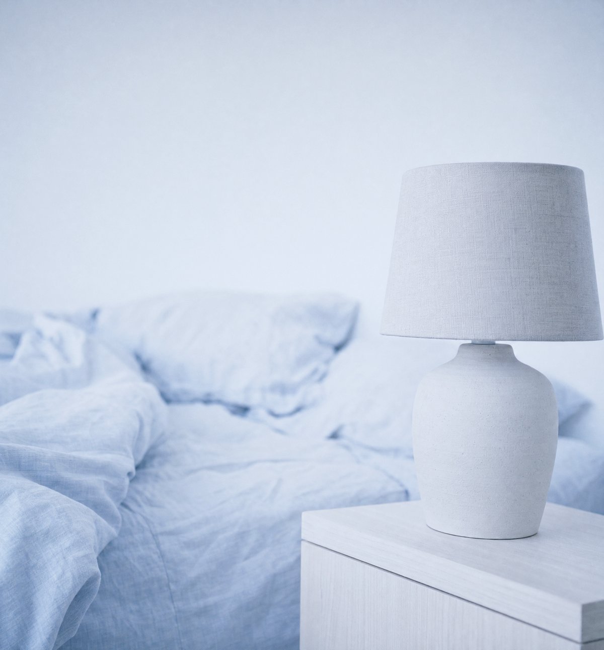 An unmade bed in pale blue linen beside a switched-off bedside lamp in early morning light