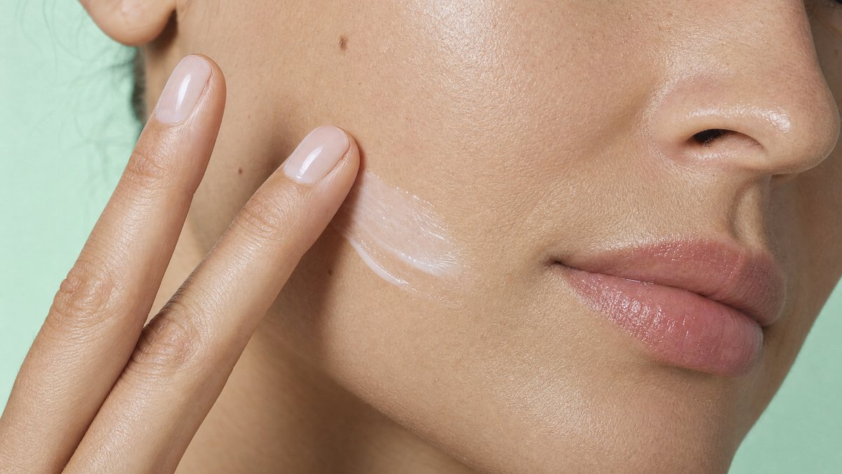 Close-up of a woman smoothing a light cream into her cheek with two fingertips