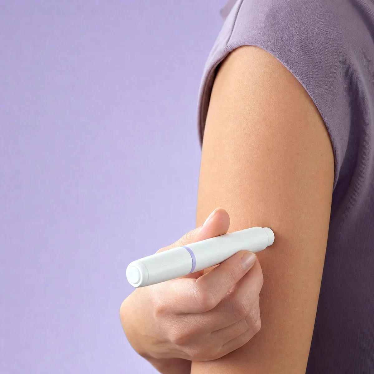 Hands holding a small unbranded auto-injector pen gently against the upper arm in soft lavender light