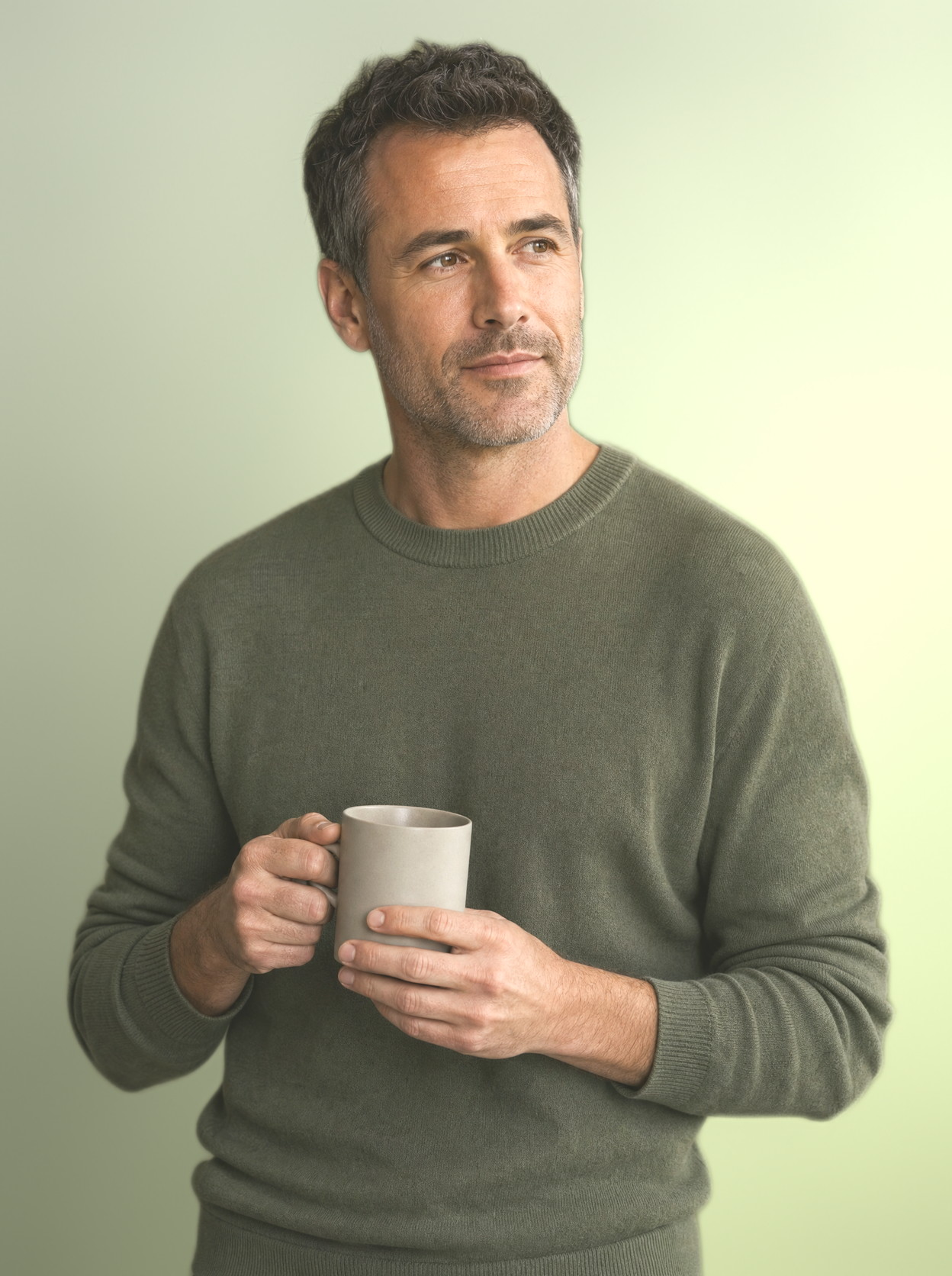 A man in his forties holding a mug of coffee on a seamless olive backdrop, calm and unhurried