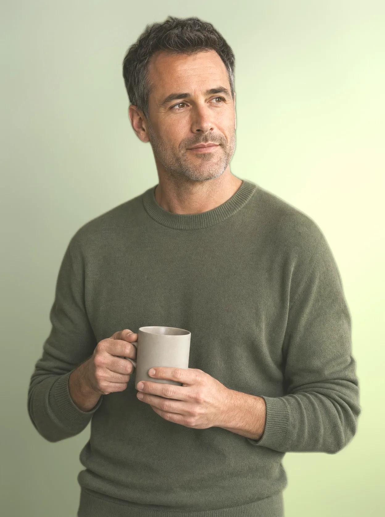 A man in his forties holding a mug of coffee on a seamless olive backdrop, calm and unhurried